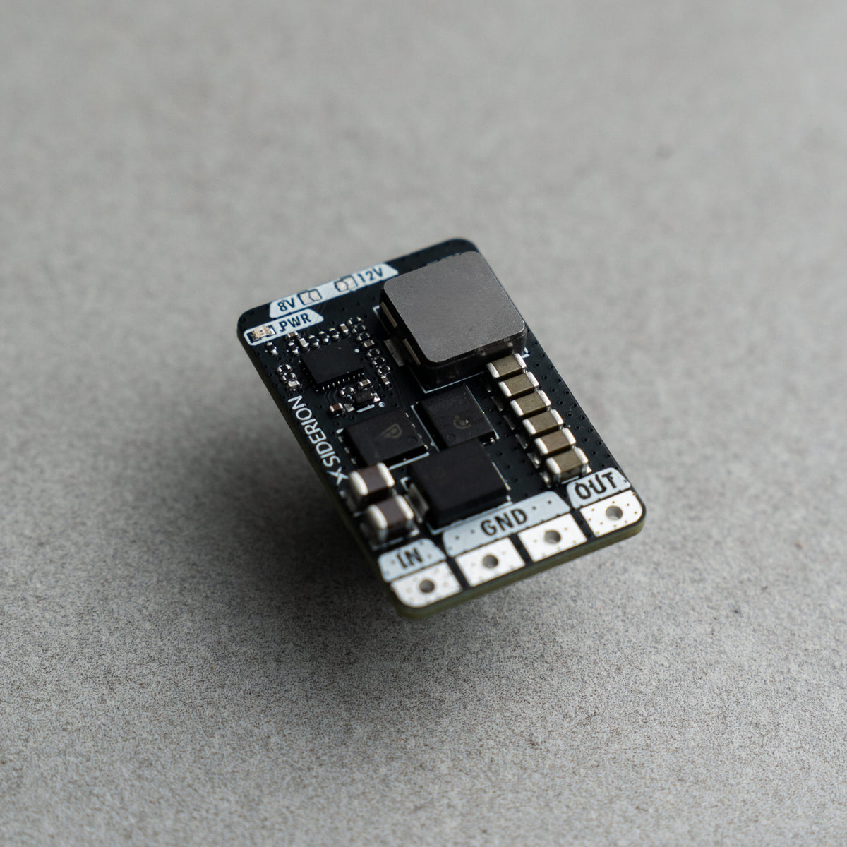 A small electronic circuit board module with a black PCB, a large black inductor, and silver terminal pads labeled IN, GND, and OUT, photographed on a textured gray surface.