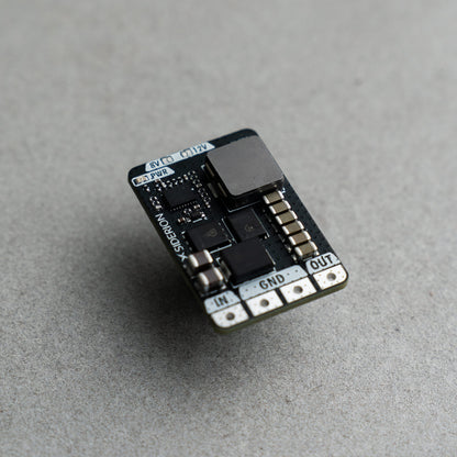 A small electronic circuit board module with a black PCB, a large black inductor, and silver terminal pads labeled IN, GND, and OUT, photographed on a textured gray surface.