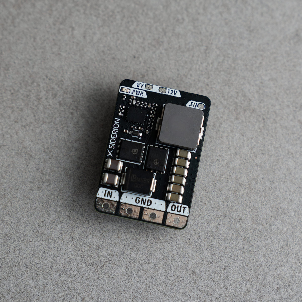 A small black electronic circuit board module with various electronic components, labeled with input/output terminals IN, GND, and OUT, resting on a textured gray surface.