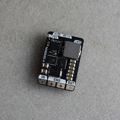 A small black electronic circuit board module with various electronic components, labeled with input/output terminals IN, GND, and OUT, resting on a textured gray surface.