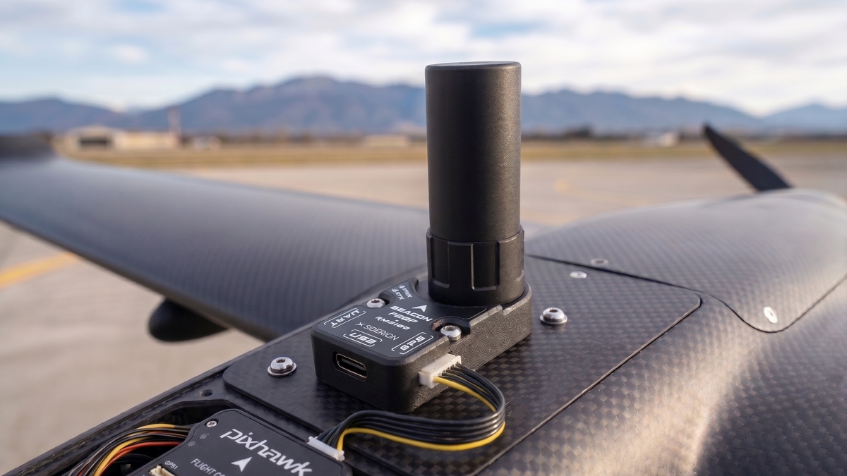A close-up of the Beacon F20P with mounted helical antenna mounted on a carbon fiber drone wing, with a Pixhawk flight controller nearby and a blurred airfield and mountains in the background.