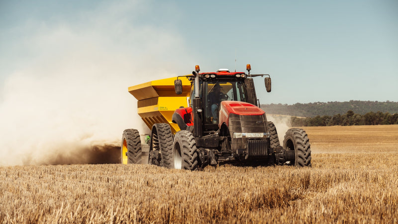 A red and black tractor pulling a yellow grain cart through a dry golden field, kicking up a large cloud of dust under a clear blue sky.