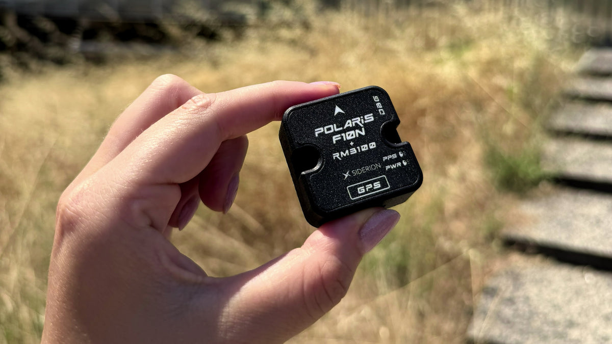 A hand holding a small black Polaris F10N GPS tracker outdoors with dry grass and stone steps blurred in the background.