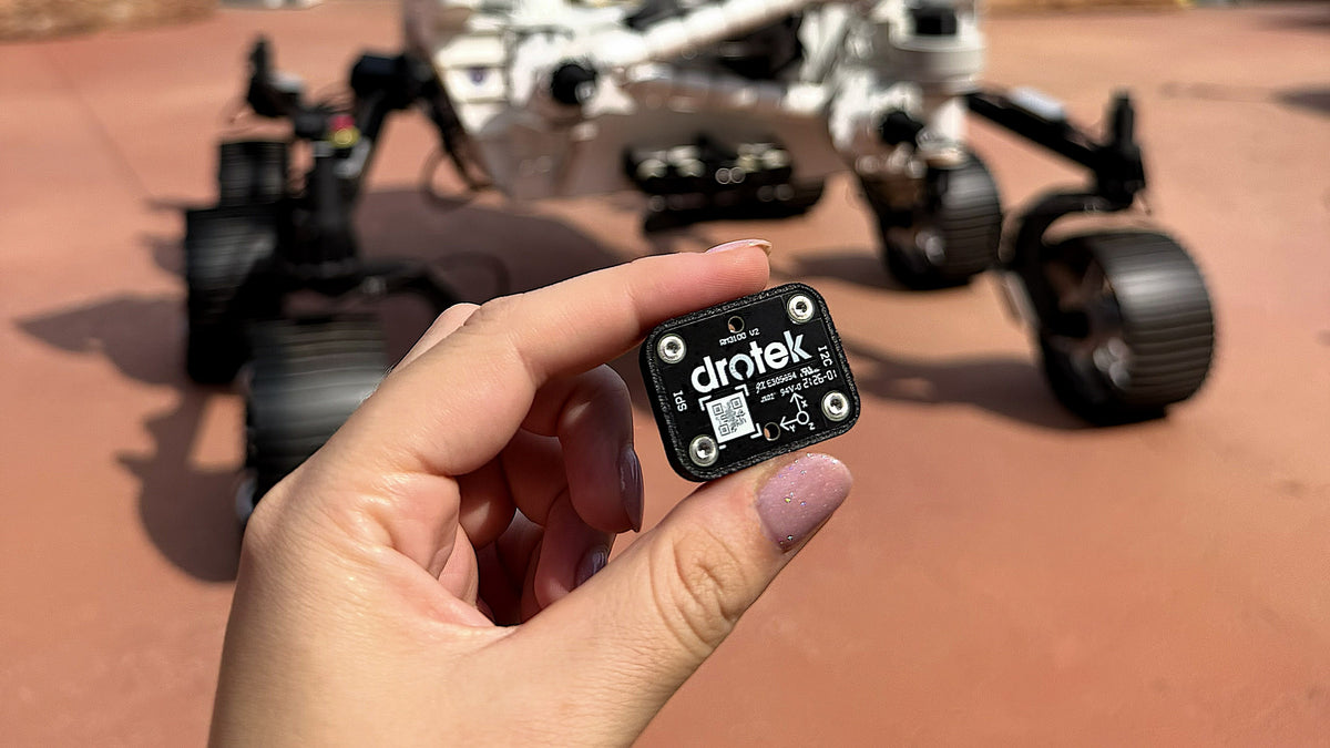 A hand holds a small black Drotek RM3100 module in the foreground, with a blurred rover or robotic vehicle in the background on a reddish outdoor surface.